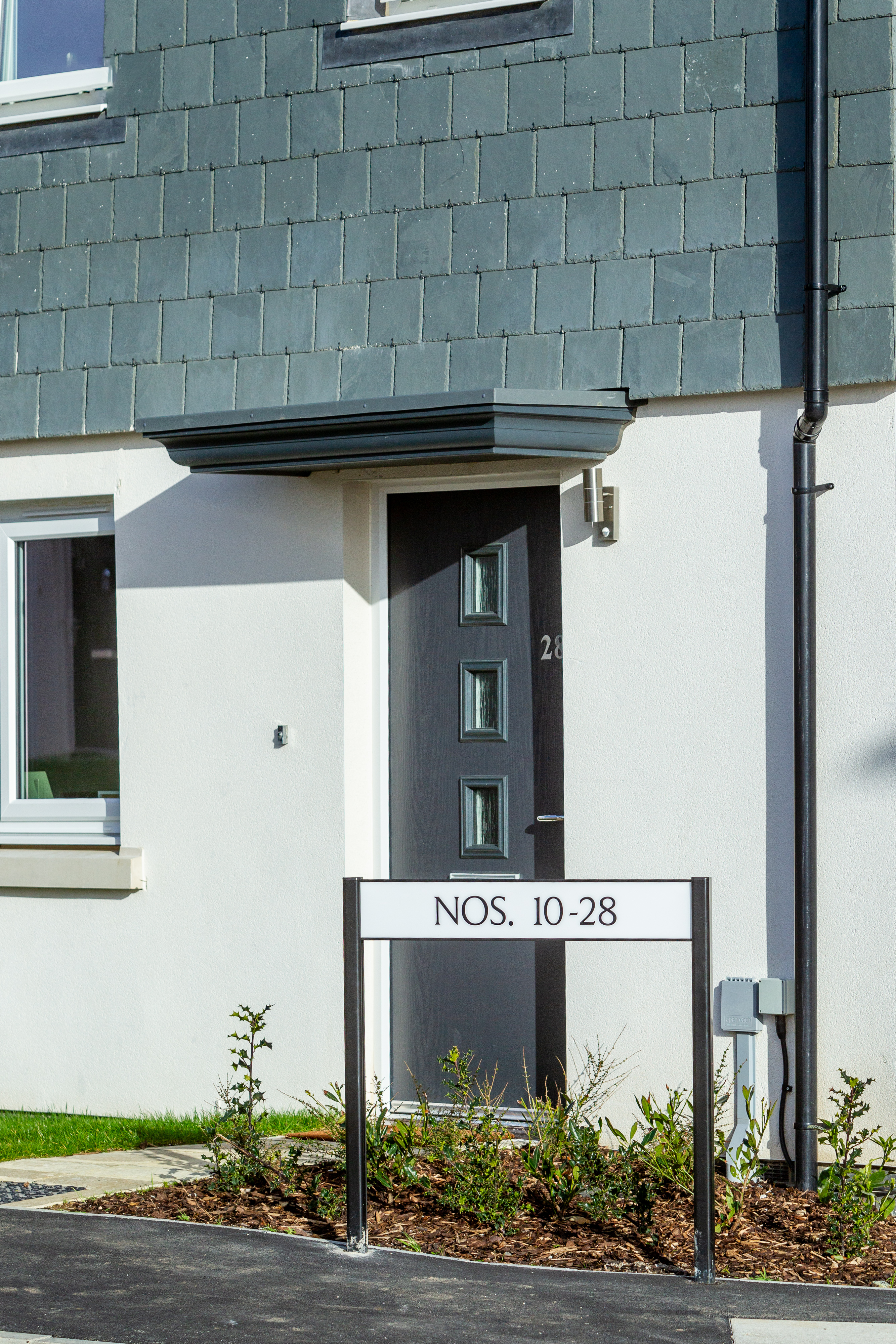 The image shows the front door of a modern house with a small garden area and a sign indicating house numbers 10 to 28.