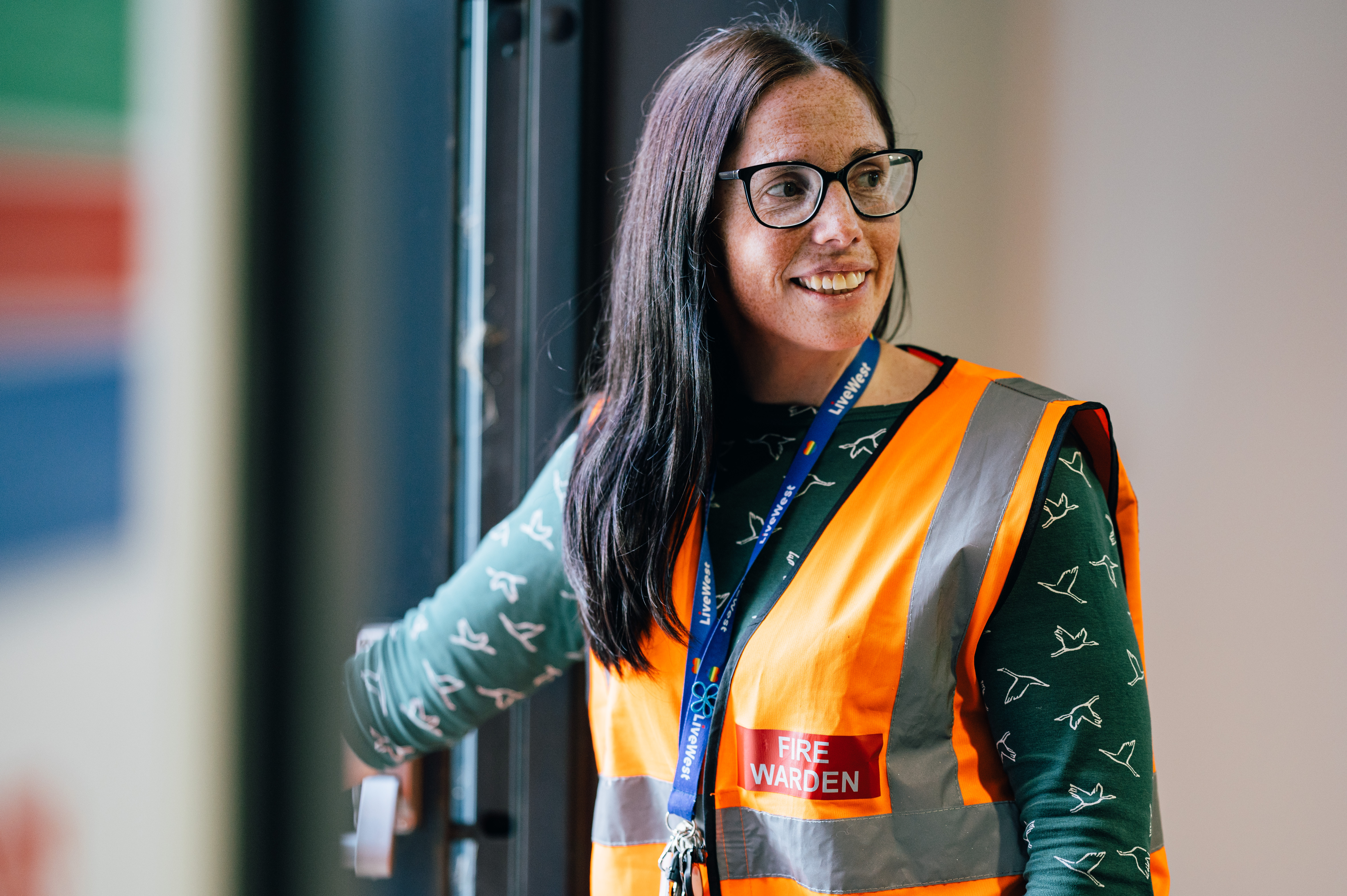 A person wearing glasses and an orange high-visibility vest labelled "Fire Warden" is standing by a door, smiling and holding the door open.