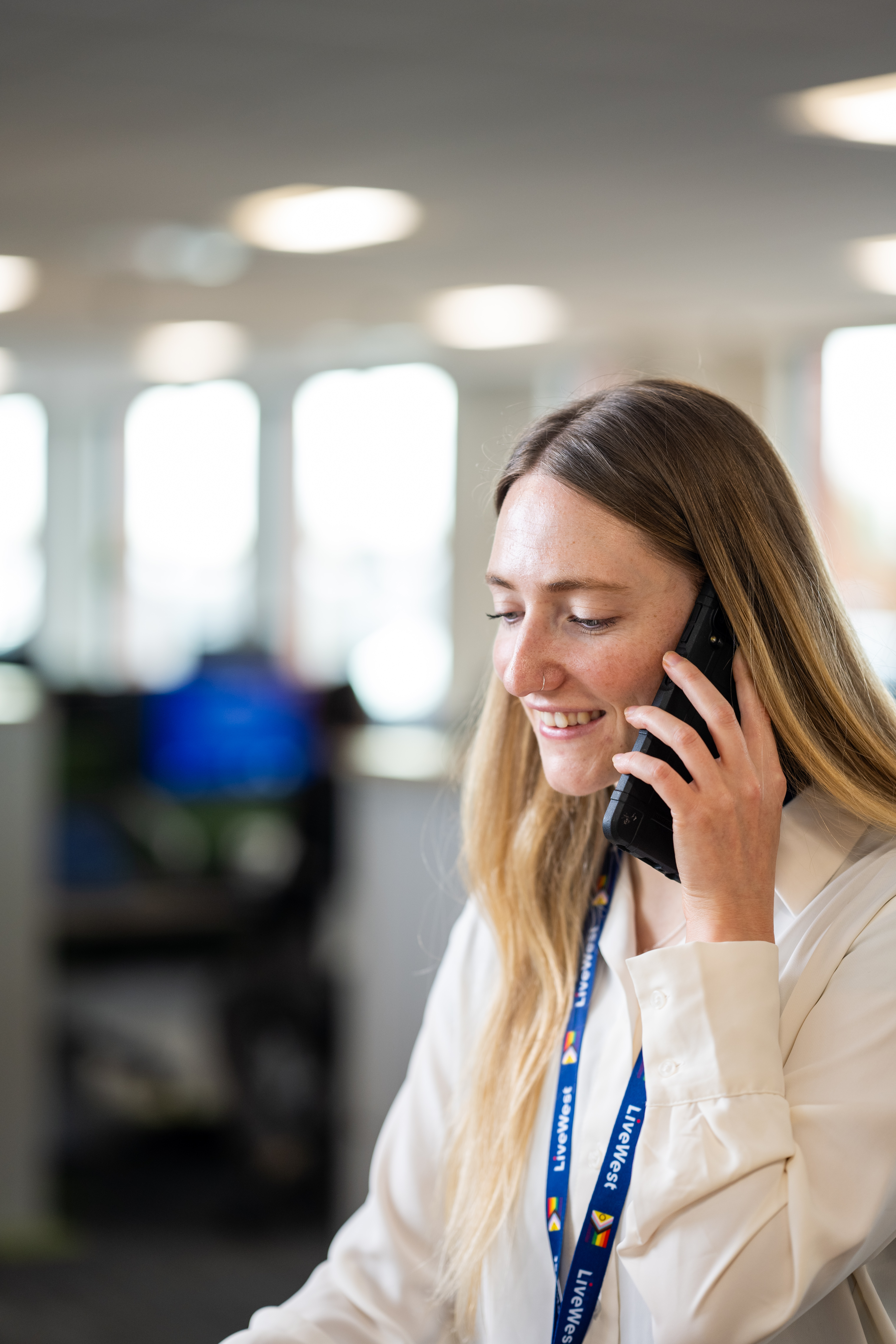 A woman is smiling while talking on a phone in an office setting.