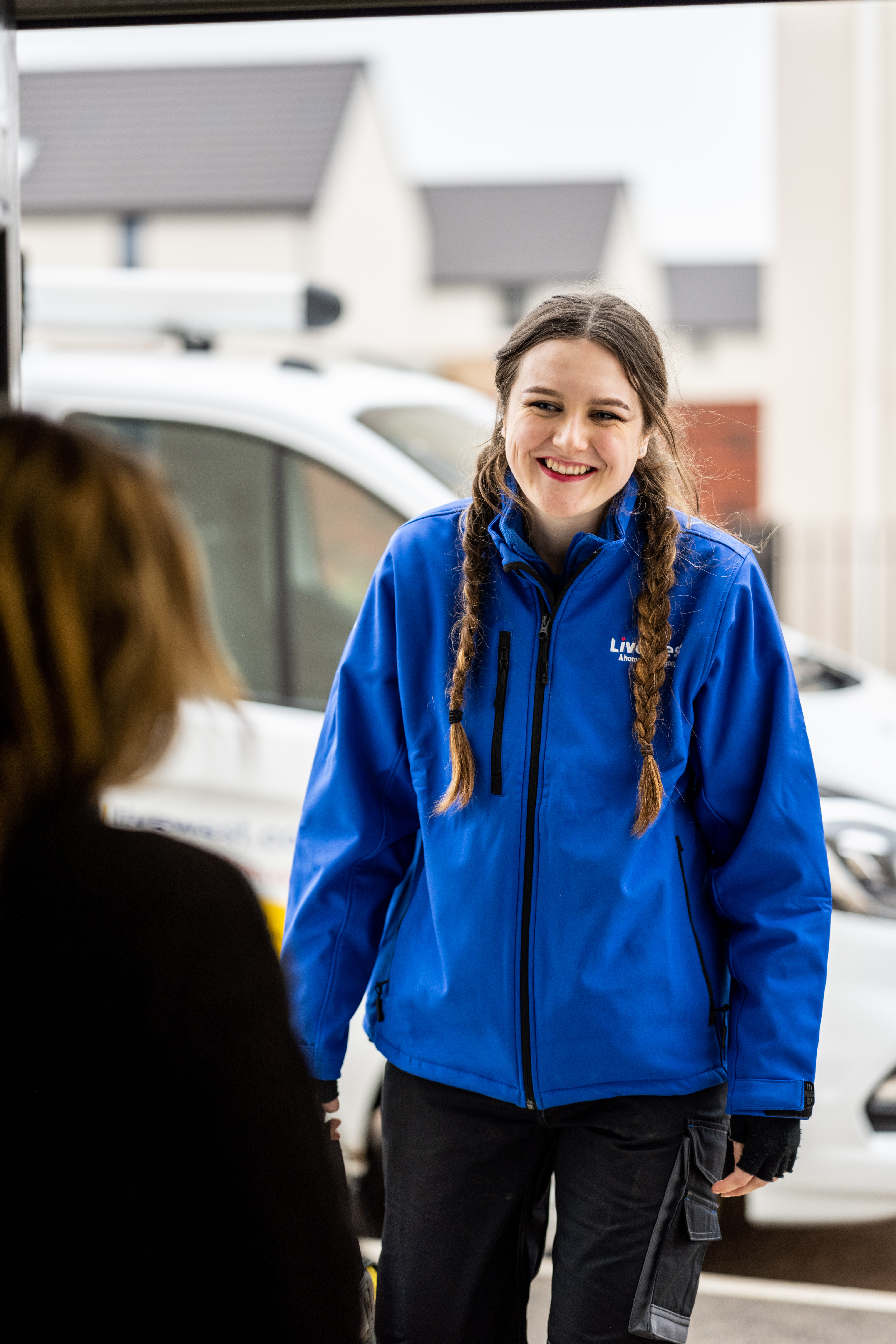 A young woman in a blue jacket is smiling and talking to another person outside in a residential area.
