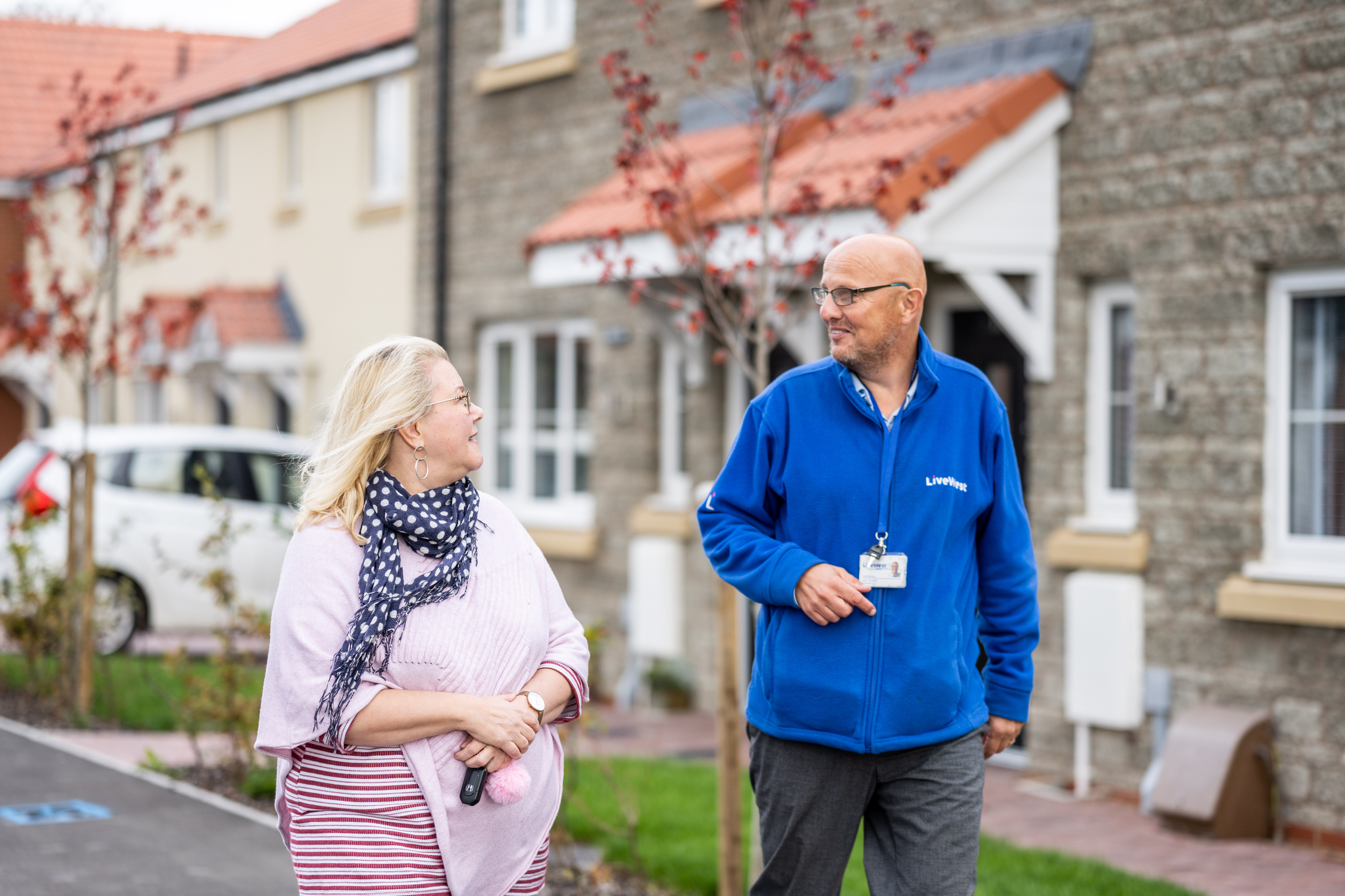 A woman and a man in a blue jacket are walking and talking on a residential street with stone houses in the background.