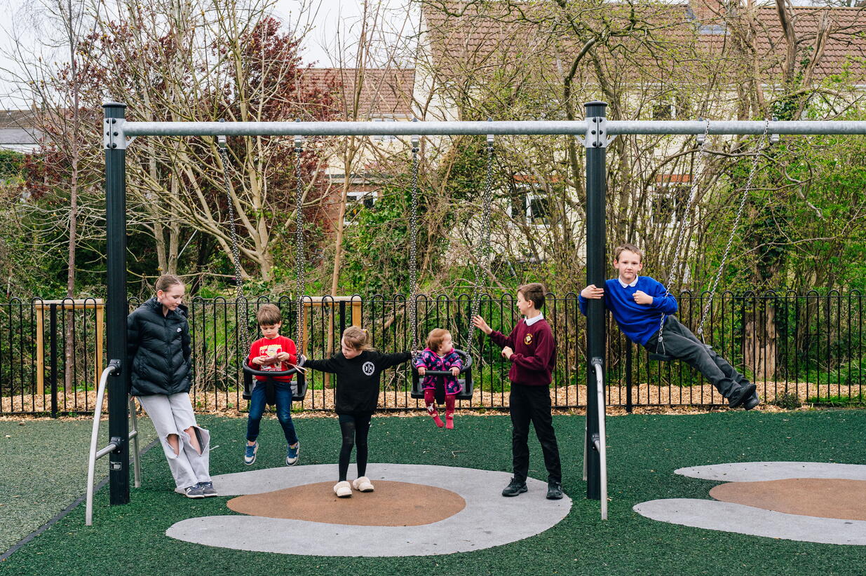 Five children are playing on and around swings in a playground, with a woman standing nearby watching them.