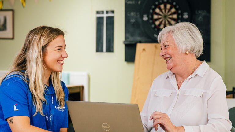 A younger woman is helping an older person use a laptop in a bright, relaxed room.