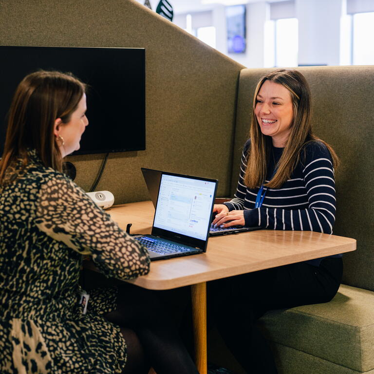 Two people are having a friendly conversation while working on laptops in a modern office space.