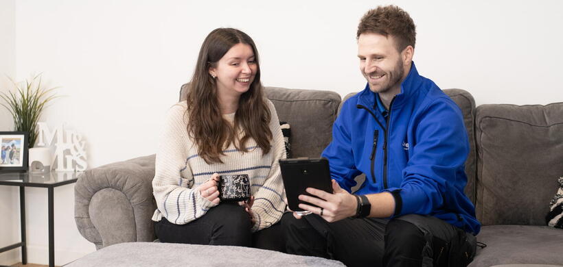 A man and a woman are sitting on a sofa in a living room, with the man holding a tablet and the woman holding a mug as they talk.