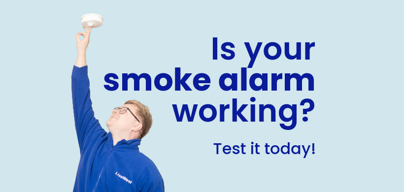 A person wearing glasses and a blue LiveWest jacket is pressing a smoke alarm on the ceiling in a kitchen.