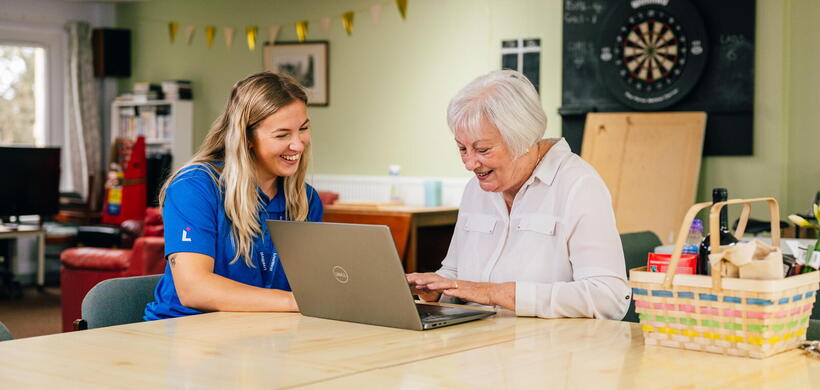 A younger woman is helping an older person use a laptop in a bright, relaxed room.