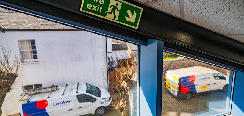 The image shows two LiveWest vans parked outside a building, viewed from inside near a fire exit sign.