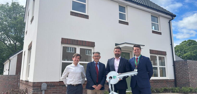 Four people stand outside a newly built two-storey house holding a large branded New Local Homes Persimmon key during a handover event.