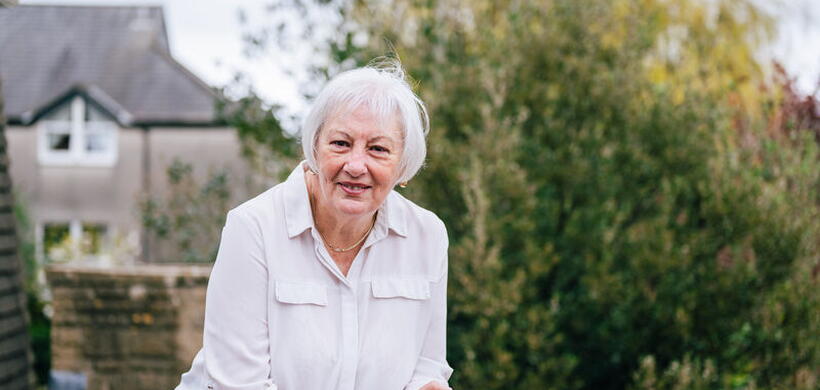 Older adult with short white hair leaning on a railing in a garden, with a house and greenery behind.