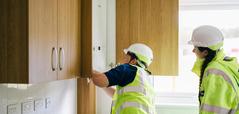 Two workers wearing hard hats and high-visibility vests are inspecting a boiler inside a kitchen cabinet.