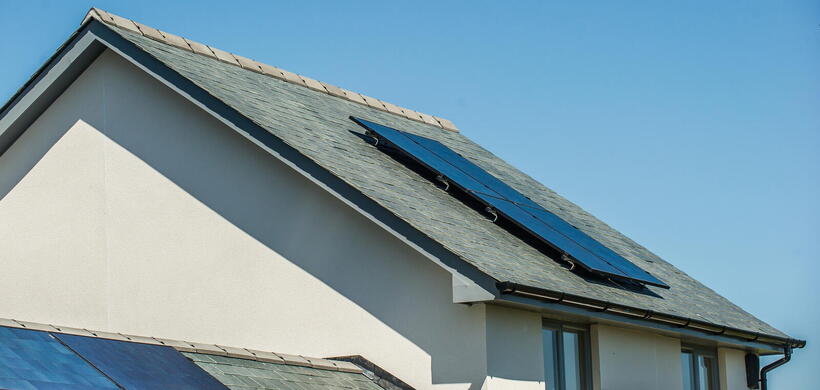 The image shows solar panels installed on the sloping roof of a white house under a clear blue sky.