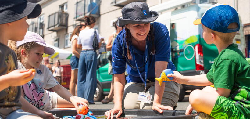 A woman in a blue shirt and sun hat plays with two children around a water tray outdoors on a sunny day.