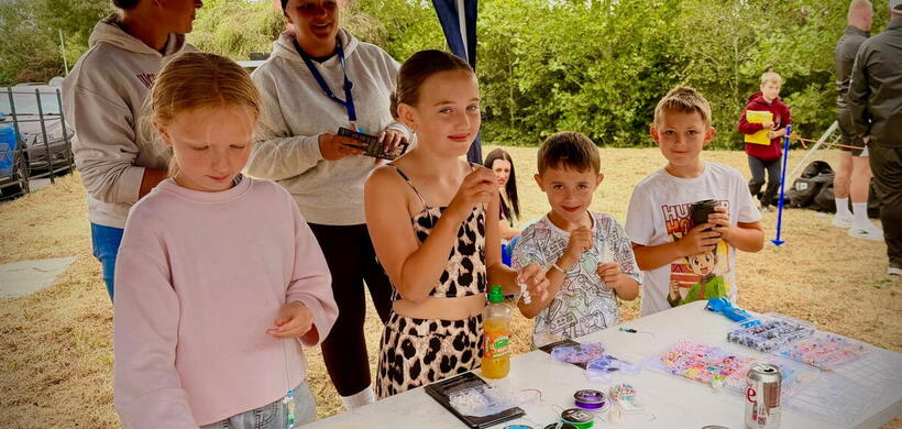 Children and adults making beaded jewellery at a table under a blue gazebo at an outdoor event.