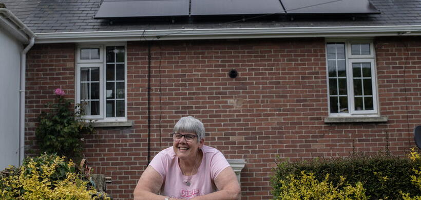 A smiling woman leaning on a garden gate in front of a house with solar panels.