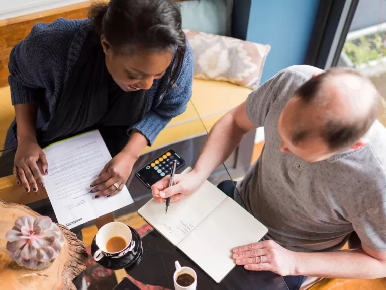 Two people sat at a table working out a budget with a calculator, pen and paper.