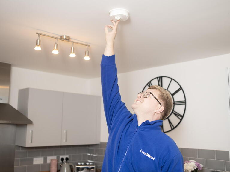 A person wearing glasses and a blue LiveWest jacket is pressing a smoke alarm on the ceiling in a kitchen.