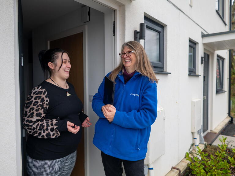 A woman wearing a blue LiveWest jacket is standing outside a house, smiling and talking with another woman who is standing in the doorway.