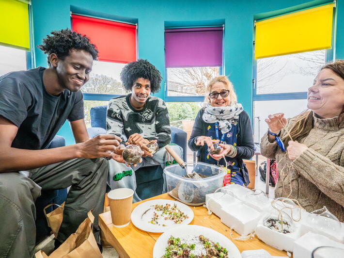 Four people sit together in a bright room, smiling and making decorations on a table.