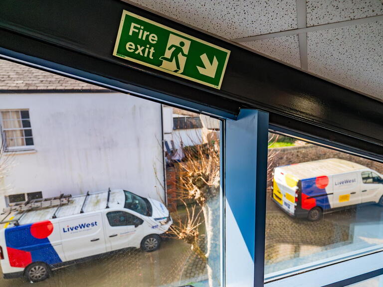The image shows two LiveWest vans parked outside a building, viewed from inside near a fire exit sign.