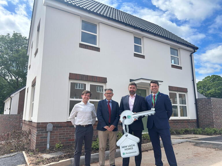 Four people stand outside a newly built two-storey house holding a large branded New Local Homes Persimmon key during a handover event.