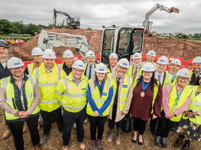 A group of people in hard hats and high-visibility vests pose together on a muddy construction site with excavators.
