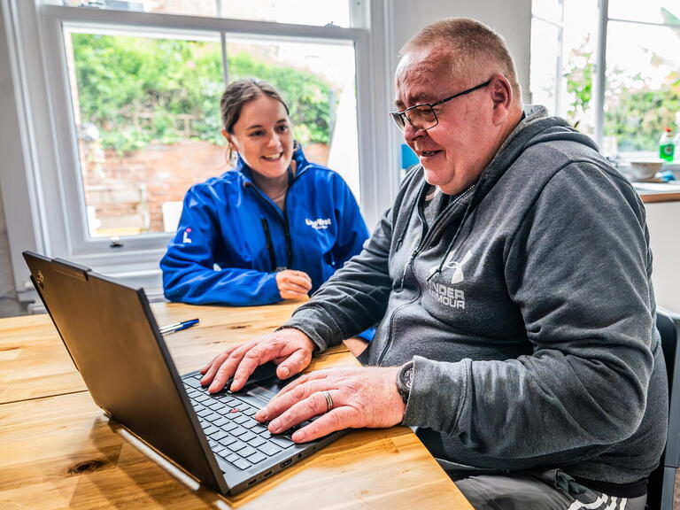 A man is using a laptop while a woman in a blue jacket watches and smiles, sitting together at a wooden table in a bright room.