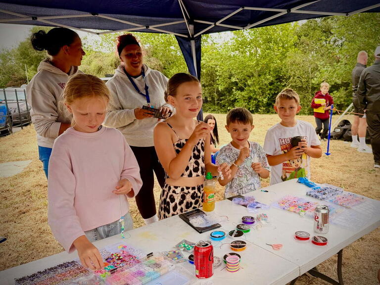 Children and adults making beaded jewellery at a table under a blue gazebo at an outdoor event.