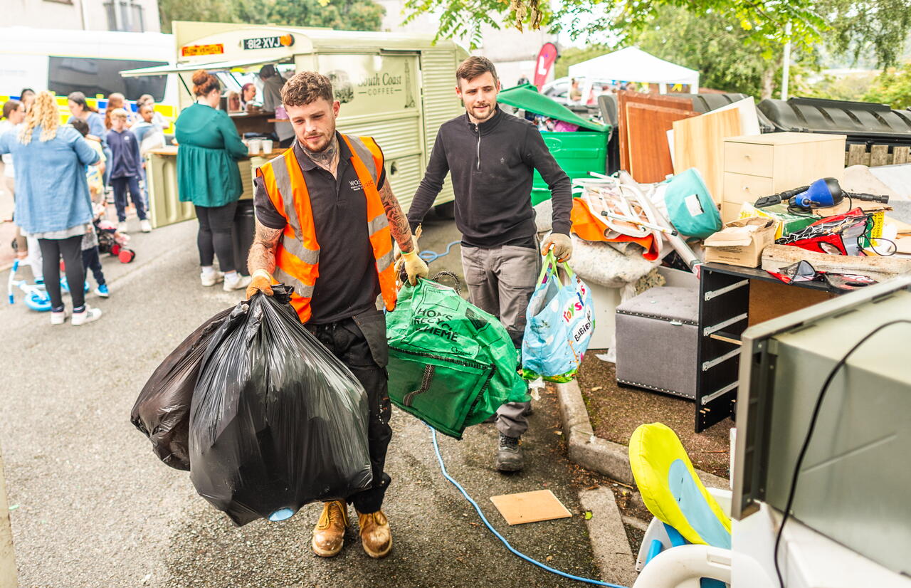 Two men carry bags and belongings at an outdoor recycling or donation event with other people and stalls in the background.