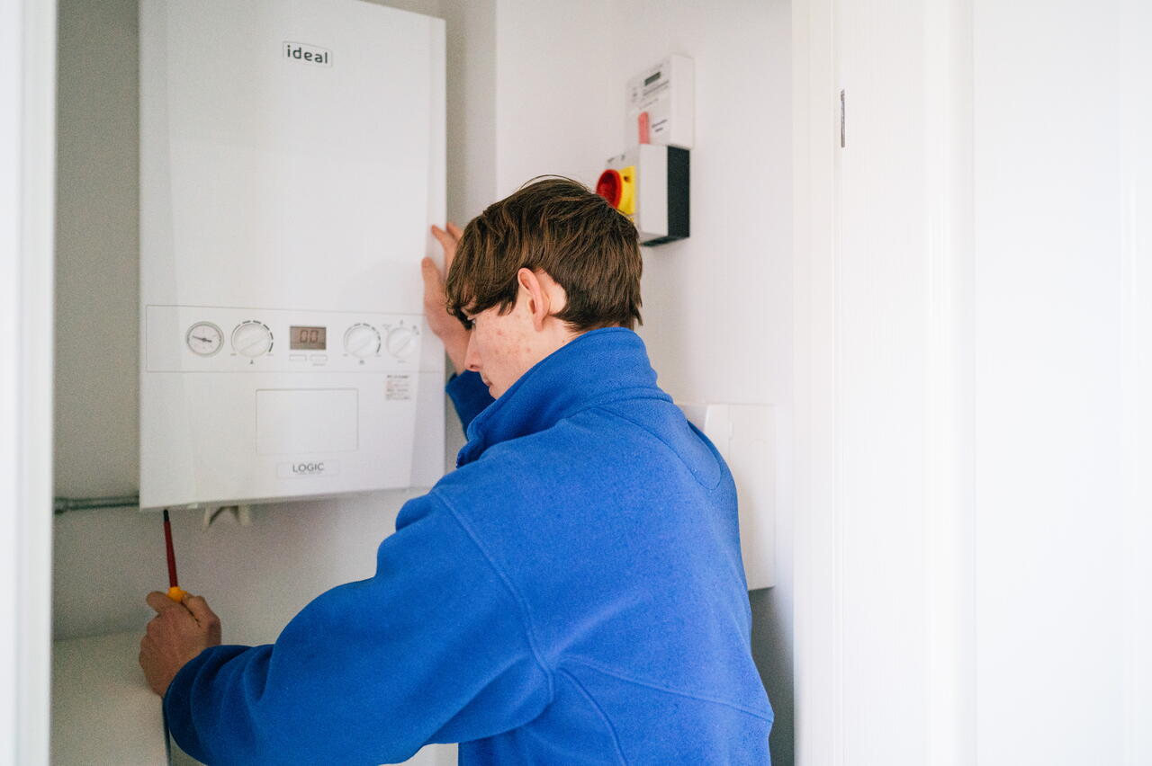 A person is fixing or checking a wall-mounted boiler in a small, white room.