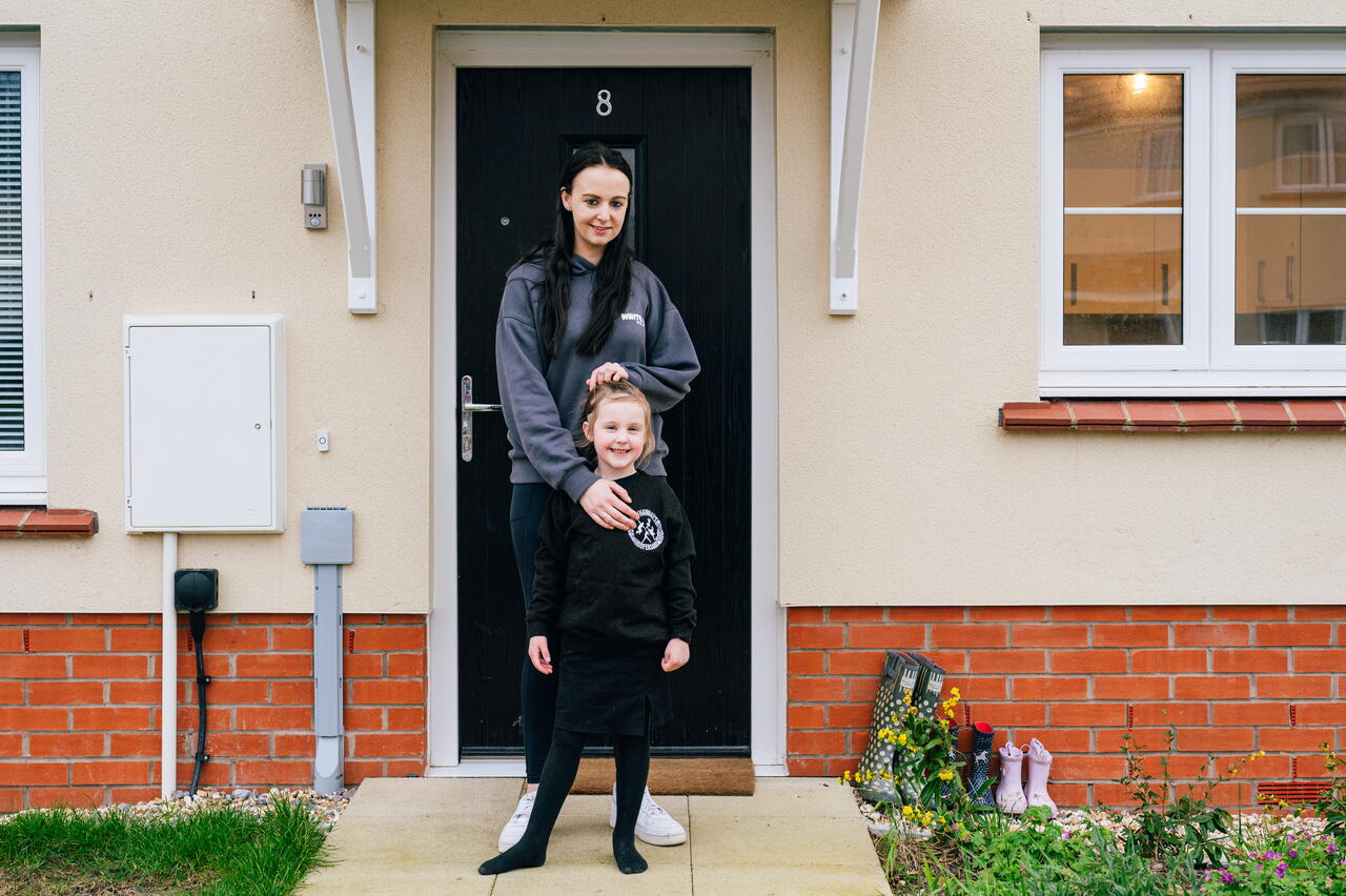 A woman stands behind a child outside the front door of a house, both smiling at the camera.