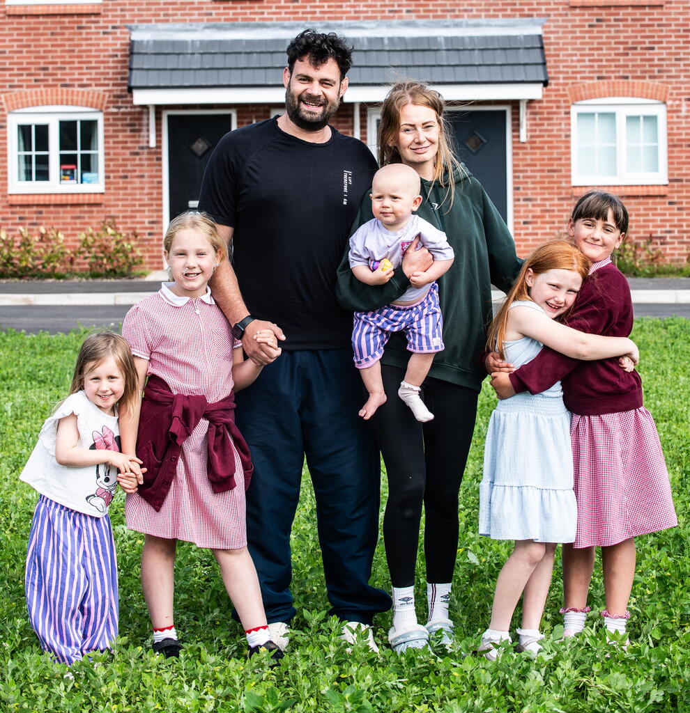A family of six stands together on the grass in front of a red brick house, smiling and holding hands.