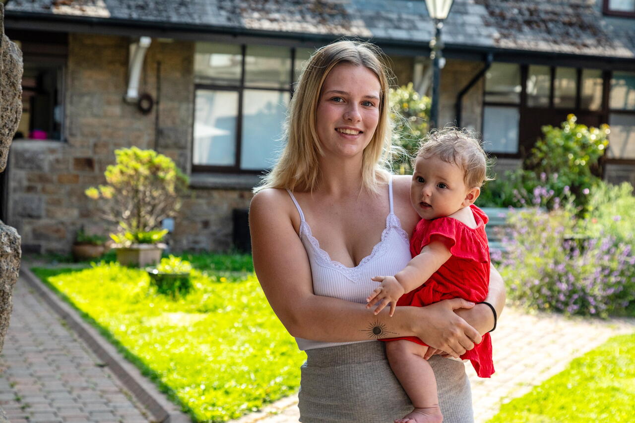 A young woman is holding a baby outside in a garden area near a house, both smiling at the camera.