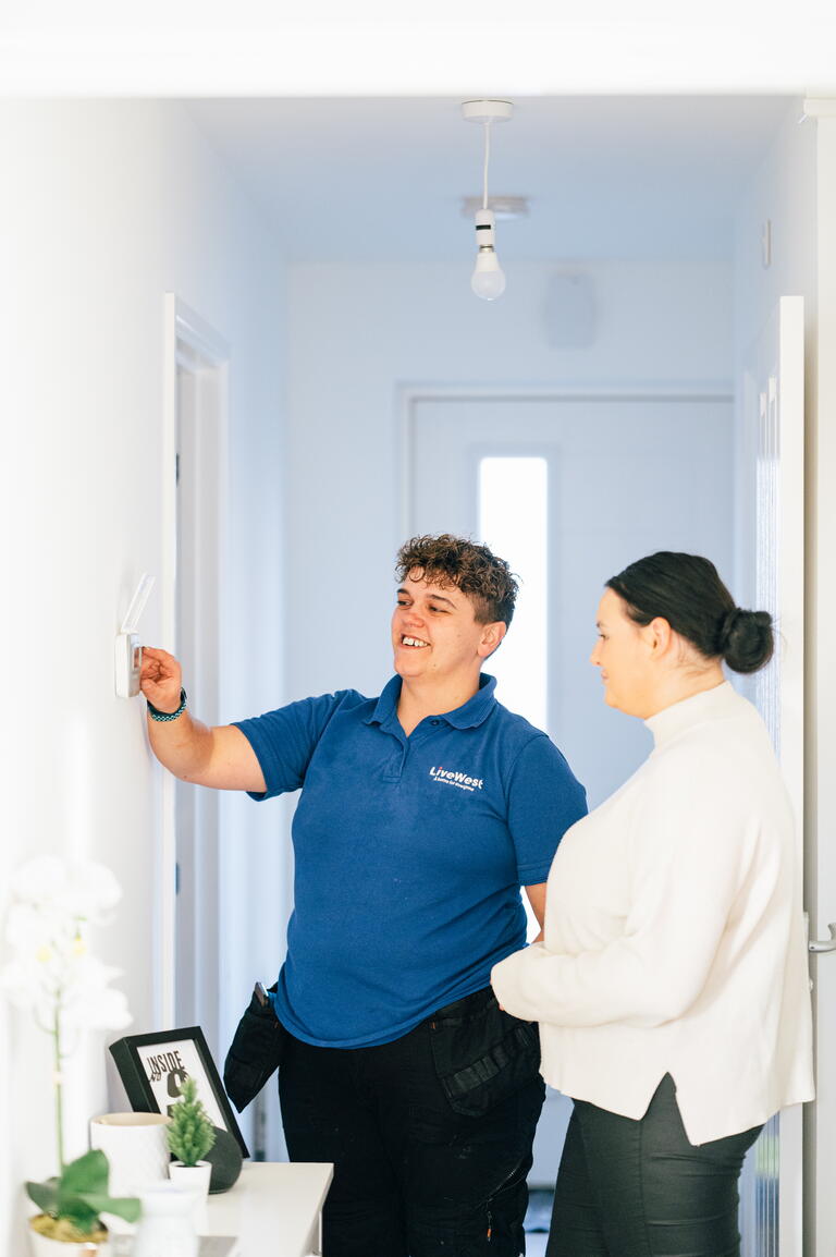 A person in a blue shirt is demonstrating how to use a light switch to another person in a white top inside a hallway.
