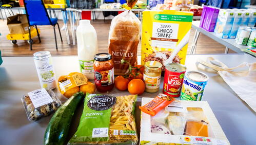 The main subject of the image is a variety of groceries arranged on a table inside a communal hall, suggesting a food distribution or collection setting.
