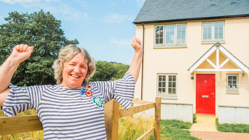 A person is smiling and raising their arms in celebration outside a house with a red door on a clear day.