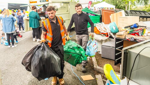 Two men carry bags and belongings at an outdoor recycling or donation event with other people and stalls in the background.