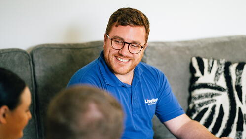 A man wearing glasses and a blue polo shirt smiles while talking to two other people in a living room.