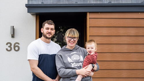 A couple is standing outside the door of their home, with one person holding a baby.