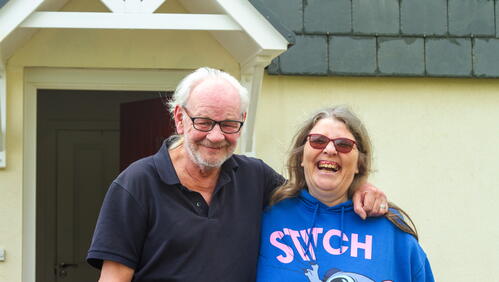 Image shows a couple smiling in front of a house.