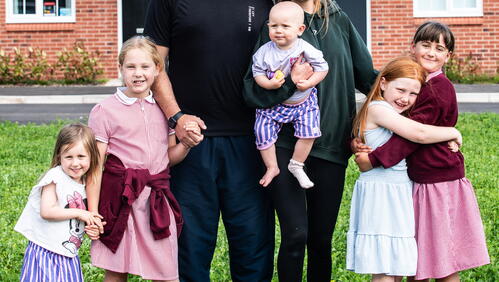 A family of six stands together on the grass in front of a red brick house, smiling and holding hands.