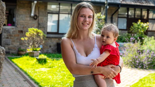 A young woman is holding a baby outside in a garden area near a house, both smiling at the camera.